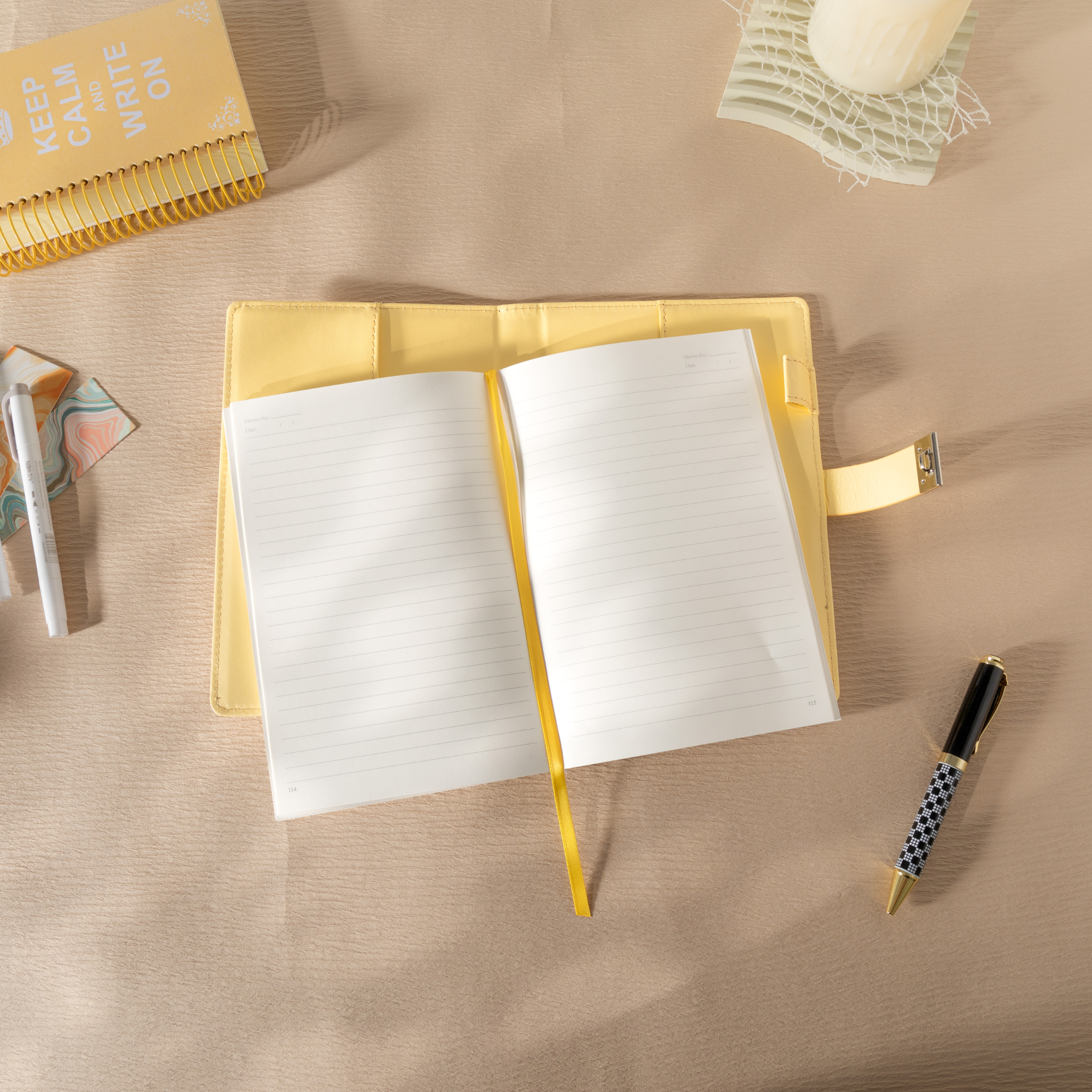 Top-down view of the open yellow vegan leather notebook lying flat on a beige desk, featuring lined paper with a yellow ribbon bookmark, an open metal lock strap, and styled with a patterned pen and a spiral notebook.