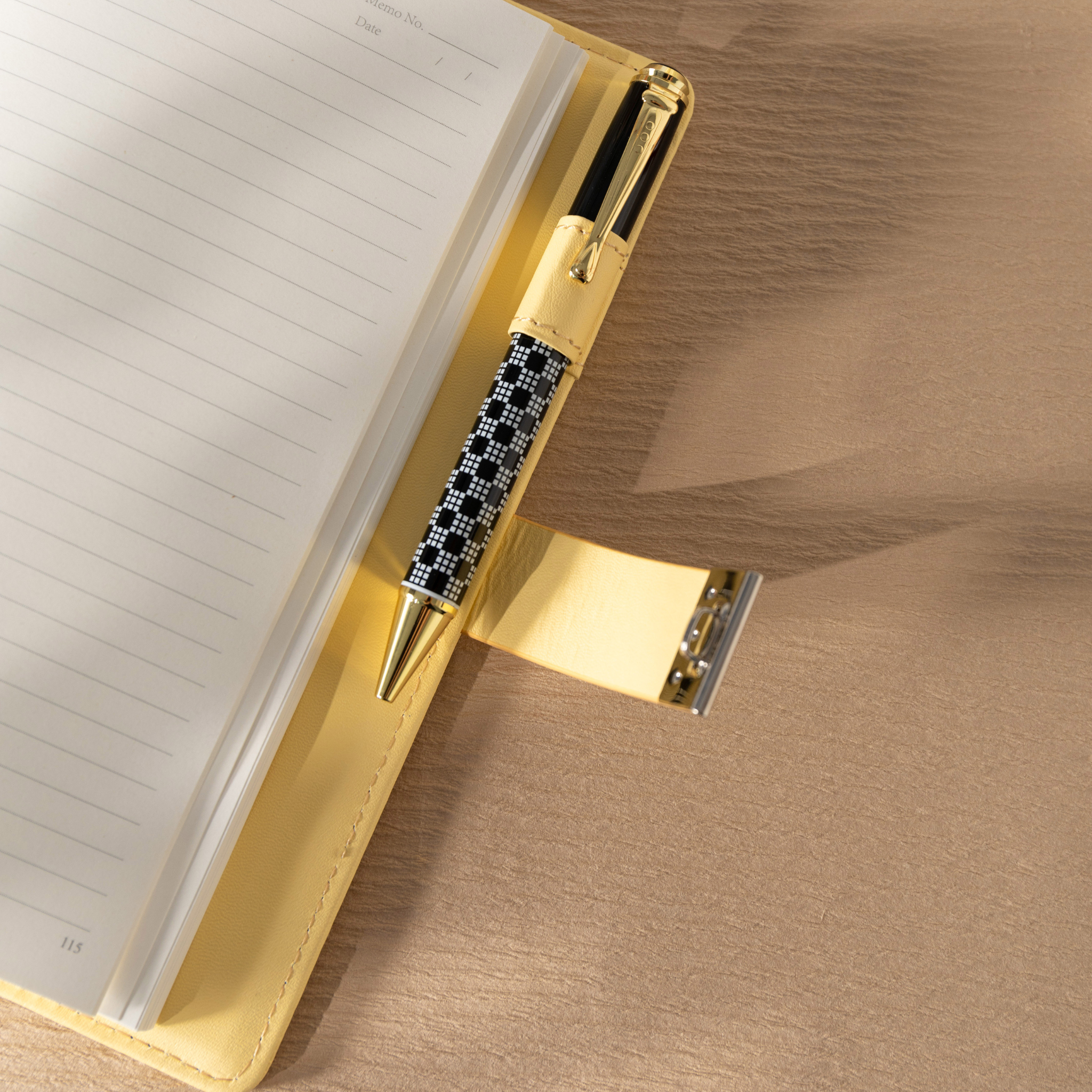 Close-up of the open yellow vegan leather notebook, showing a black and white patterned pen tucked into the side pen loop, with the metal lock strap opened and resting on the cover.