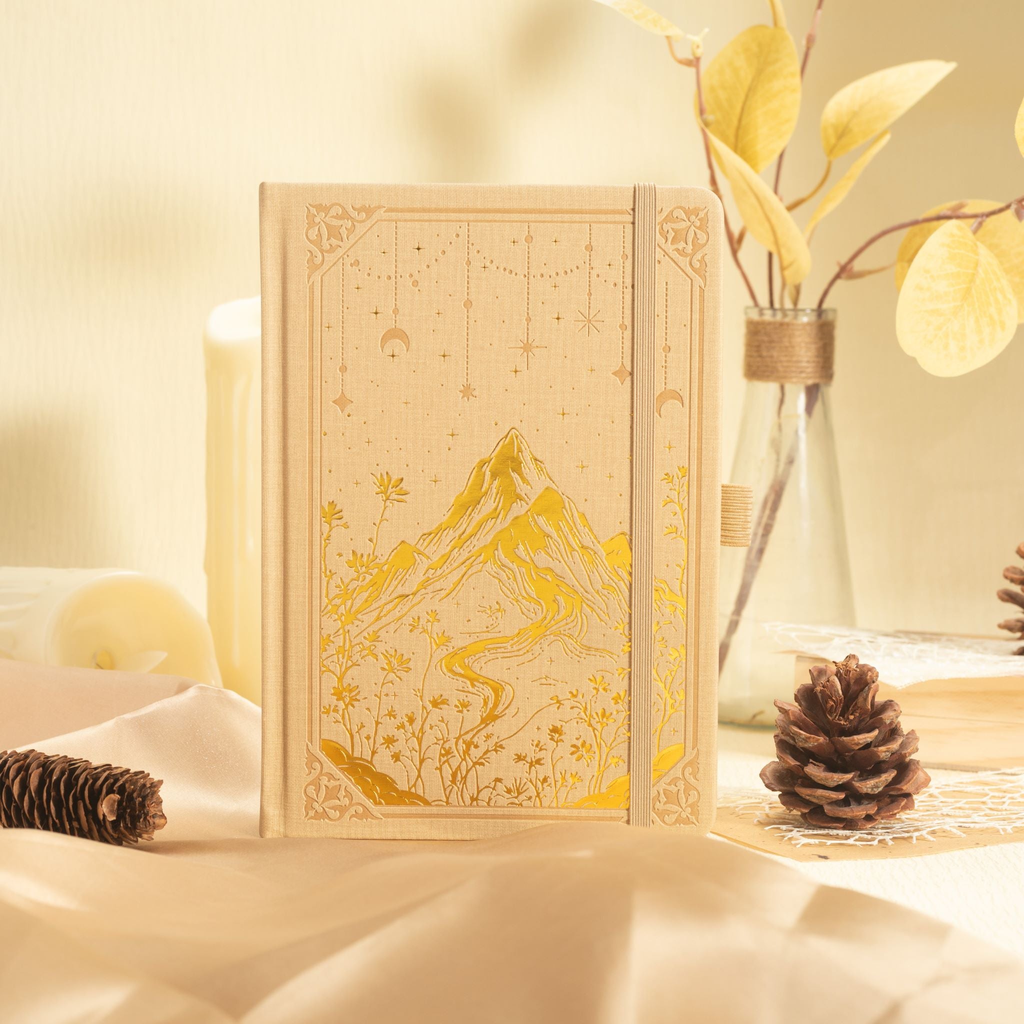 A5 beige mountain notebook standing upright in a cozy desk setup with dried flowers, highlighting the gold foil cover details.
