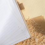Close-up of the open beige vegan leather notebook, highlighting the 100gsm lined paper interior with elegant gold borders, corner page numbers, and a matching beige elastic band.