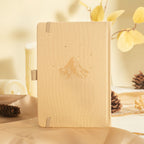Beige vegan leather notebook standing upright on a warm wooden desk, showcasing a subtle embossed mountain design and matching elastic band, surrounded by pinecones and dried leaves.
