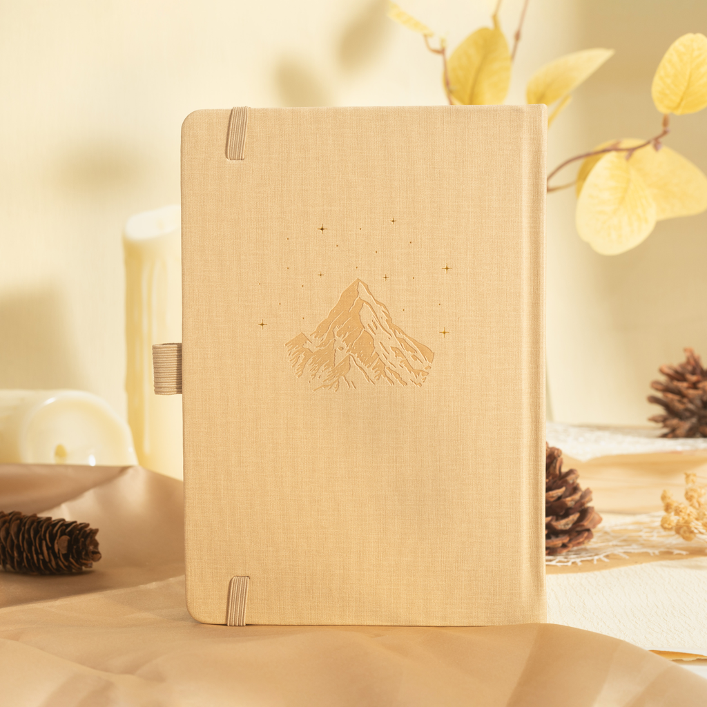 Beige vegan leather notebook standing upright on a warm wooden desk, showcasing a subtle embossed mountain design and matching elastic band, surrounded by pinecones and dried leaves.