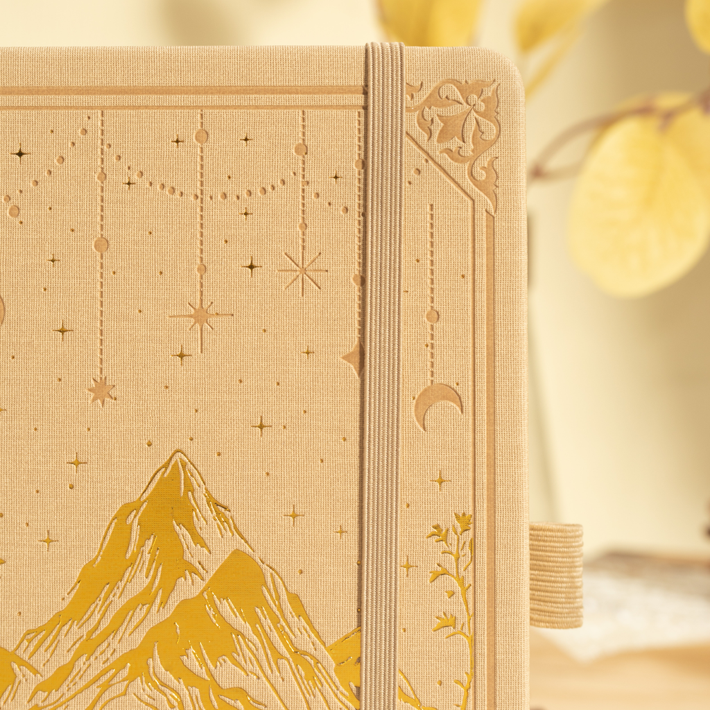 Close-up of the beige vegan leather notebook cover showing gold foil mountain and star embossing, intricate border details, and a matching beige elastic band.