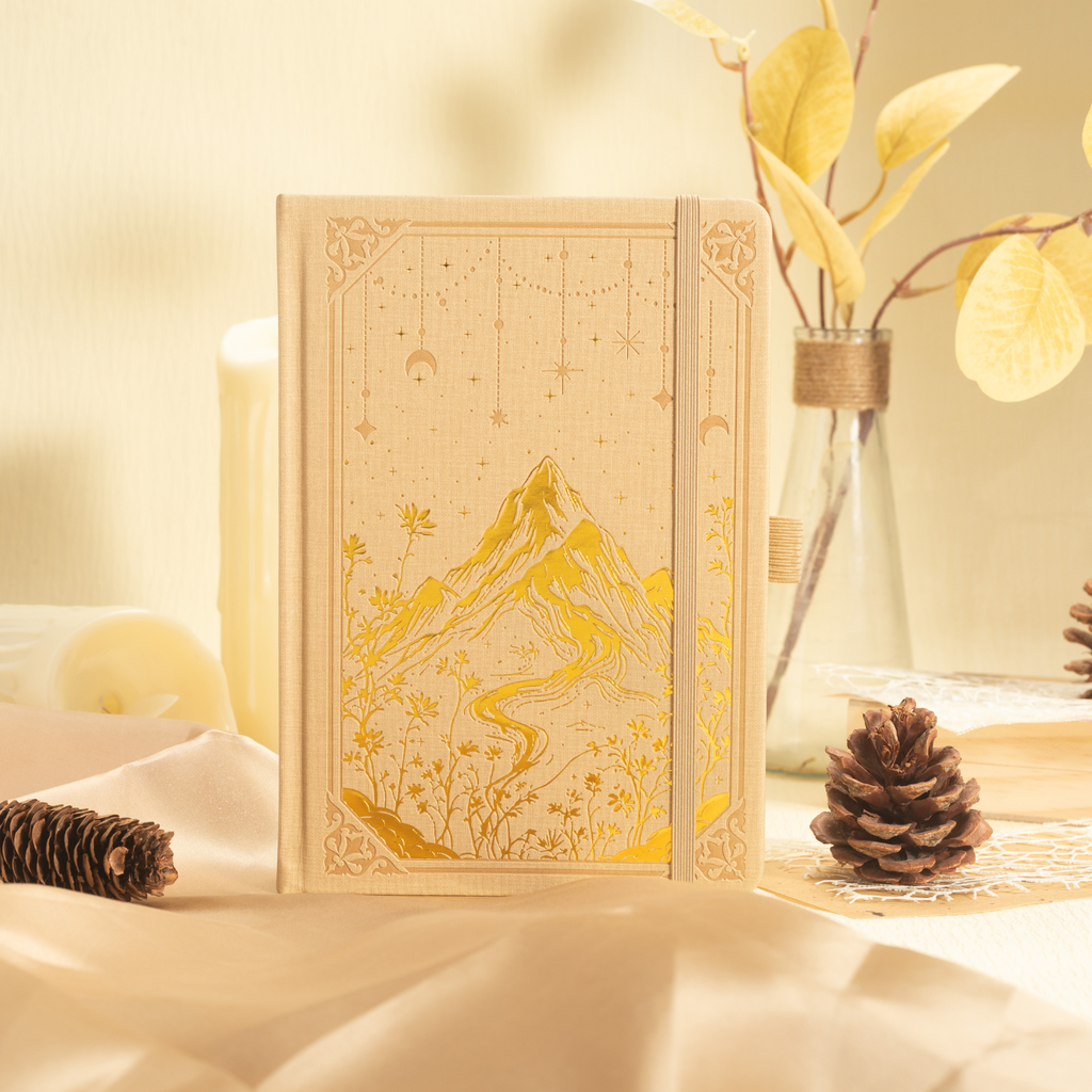 A5 beige mountain notebook standing upright in a cozy desk setup with dried flowers, highlighting the gold foil cover details.