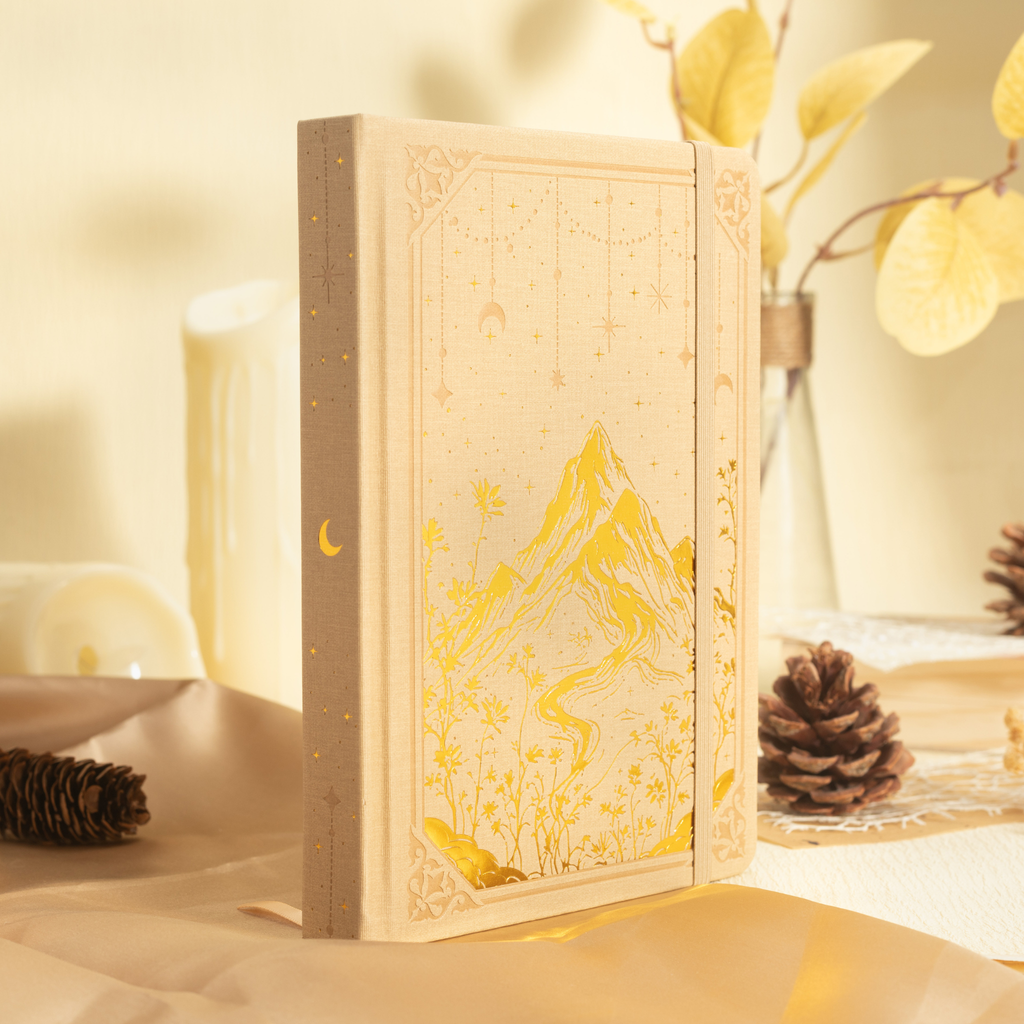 Beige A5 journal standing sideways on a desk, displaying its gold gilded page edges and a gold foil mountain cover in a warm floral setting.