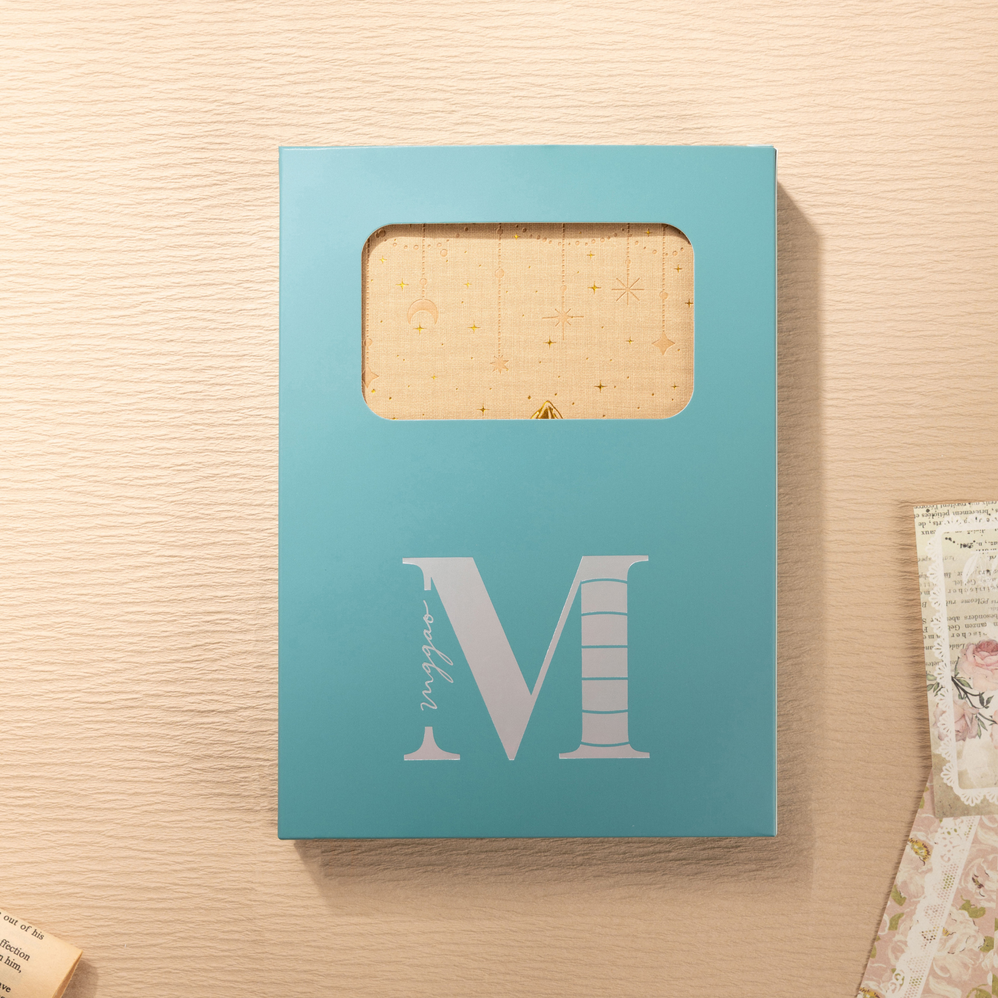 Light blue window-cut gift box featuring a white "M" logo, displaying the beige mountain vegan leather notebook inside through the cut-out.