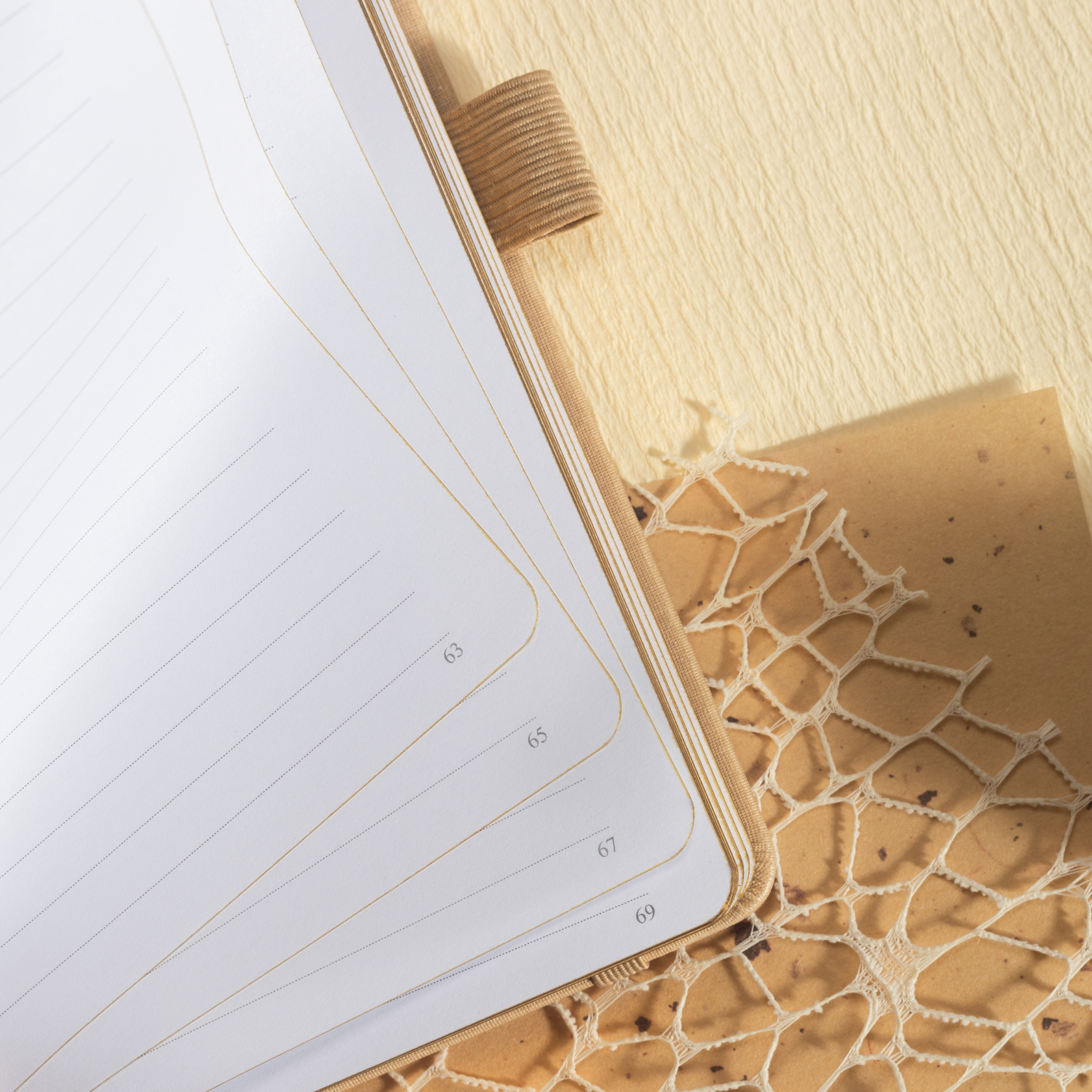 Close-up of the open beige vegan leather notebook, highlighting the 100gsm lined paper interior with elegant gold borders, corner page numbers, and a matching beige elastic band.