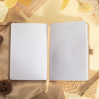 Open A5 notebook lying flat, showing the 100gsm lined paper interior with date and memo headers, a beige ribbon bookmark, and elegant gold-edged pages, styled with pinecones and a gold pen.