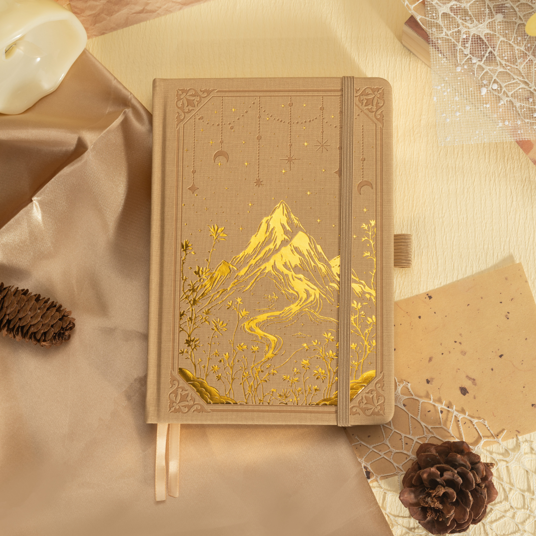 Beige vegan leather notebook lying flat on a warm fabric surface, showcasing a striking gold foil mountain and starry night cover design, with a matching beige elastic band and ribbon bookmark.