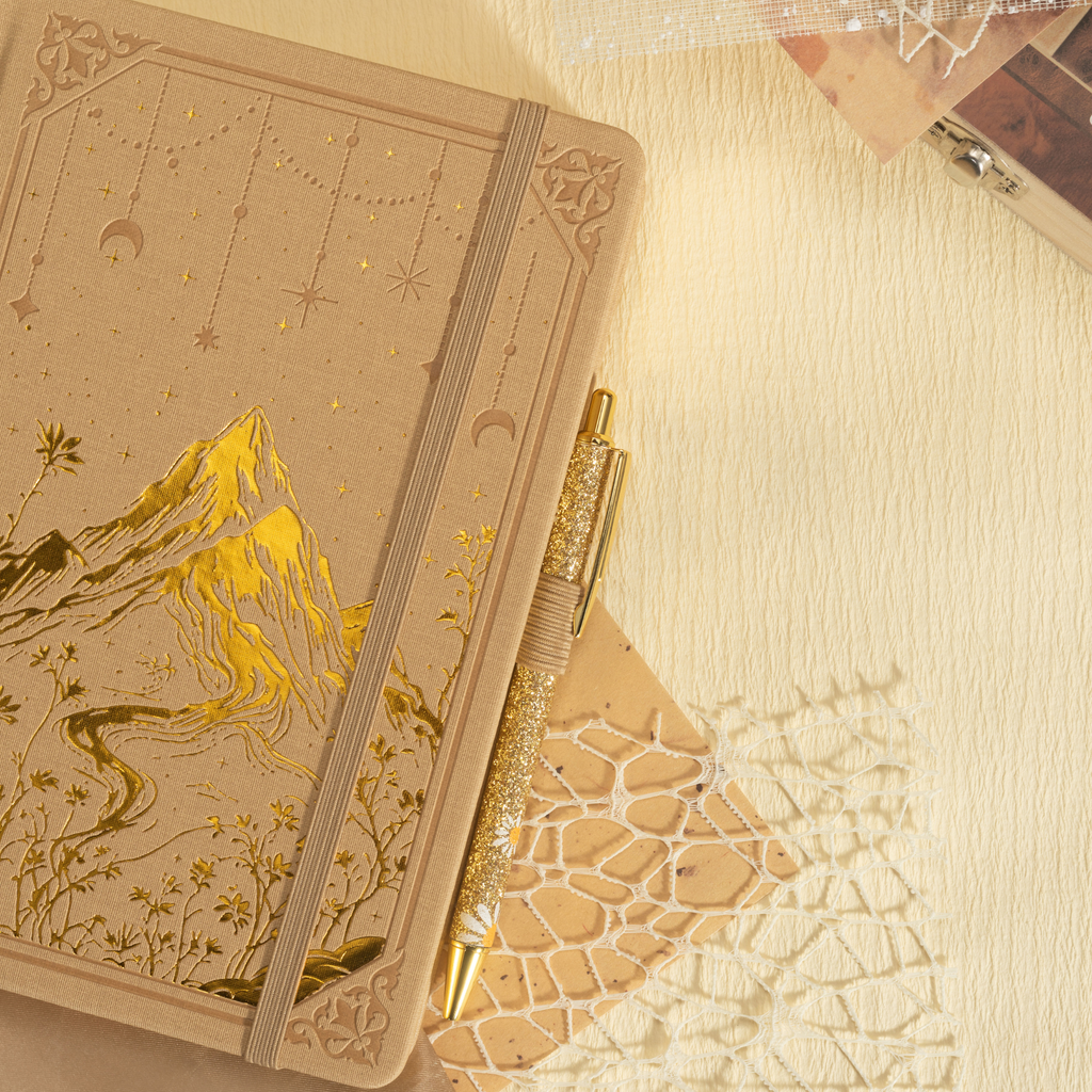Beige vegan leather journal laid flat with a matching pen and dried flowers, showcasing the gold foil mountain cover design.