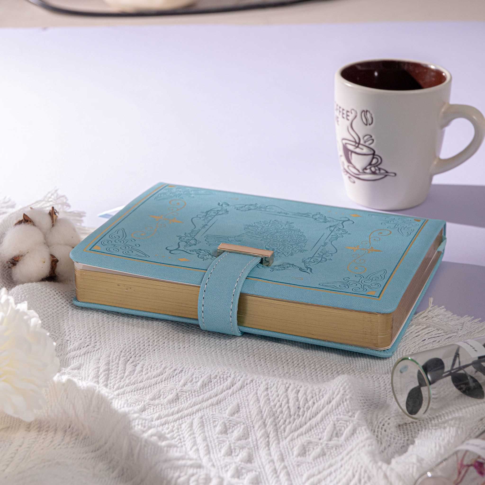 B6 Light Blue Rose Embossed Vegan Leather Journal with Gold Gilded Edges, Desk Background with Coffee Cup