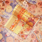 Orange, red, and purple blossom washi tapes partially unrolled on a warm floral background, showcasing intricate gold foil geometric lattice, fan-shaped floral patterns, and colorful floral motifs.