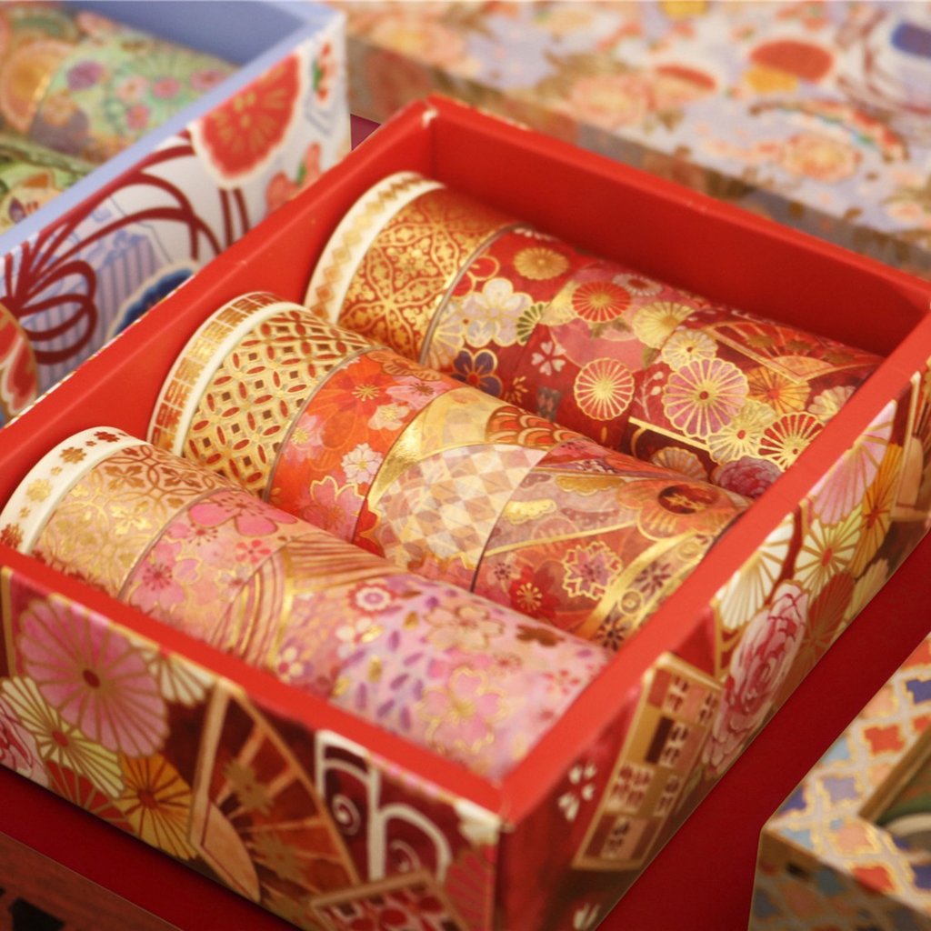 Close-up of a red floral gift box filled with multiple rolls of blossom gold foil washi tape in red, orange, and pink tones, with a blue box partially visible in the background.