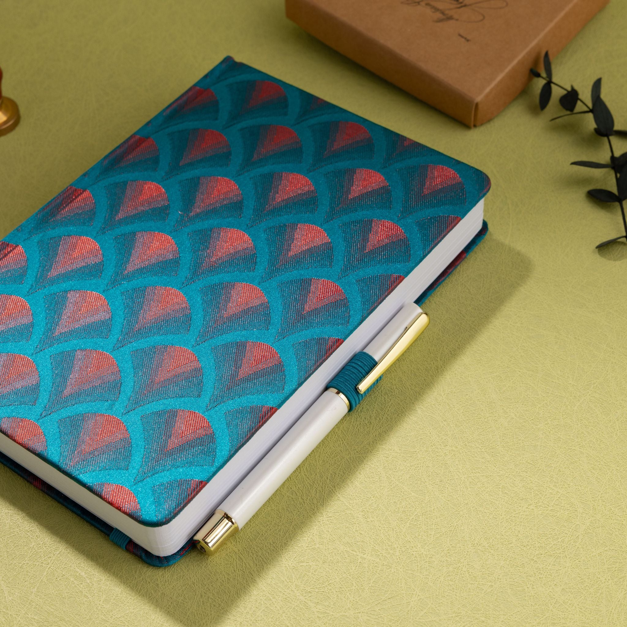 Teal-blue and red-copper embroidered scalloped fabric notebook with elastic band, holding a white and gold pen