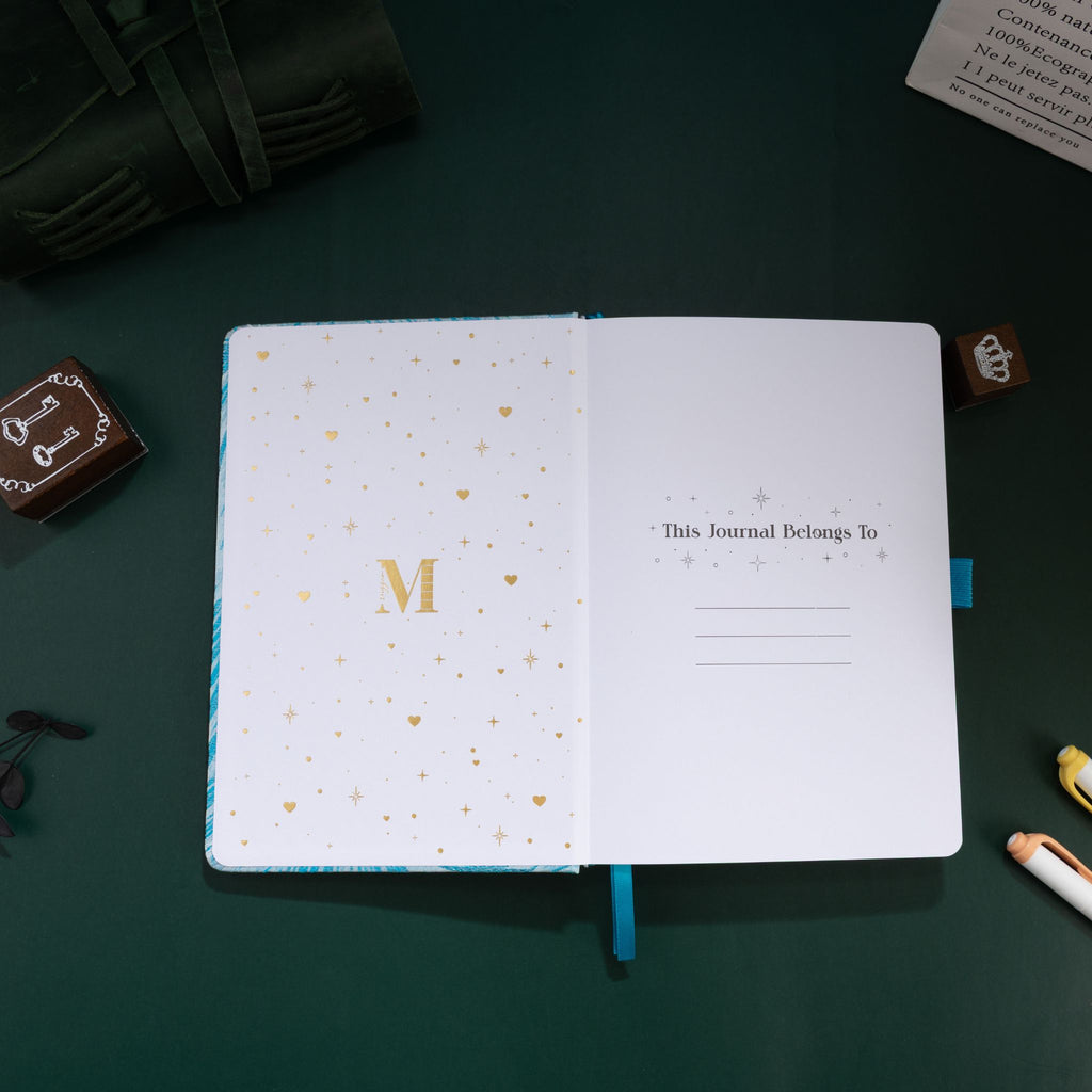 Inside title page featuring a gold foil 'M' logo and 'This Journal Belongs To' section on a light blue embroidered journal