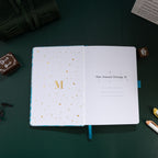 Inside title page featuring a gold foil 'M' logo and 'This Journal Belongs To' section on a light blue embroidered journal