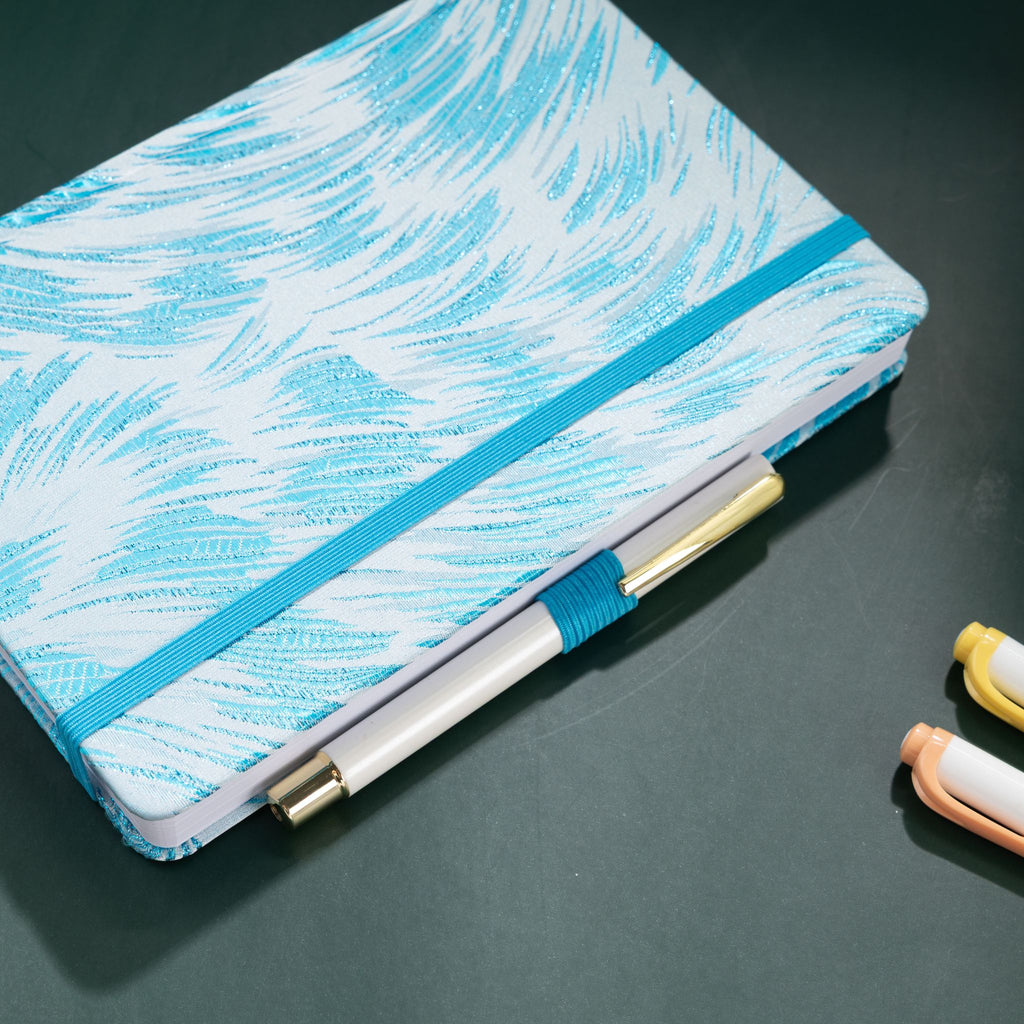 Light blue and white feather-embroidered fabric notebook with elastic band, holding a white and gold pen