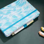 Light blue and white feather-embroidered fabric notebook with elastic band, holding a white and gold pen