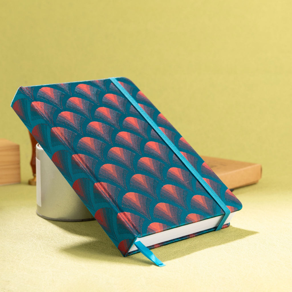 A5 teal and red embroidered scalloped fabric notebook standing tilted on a yellow desk with stationery