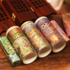 Gold foil washi tape rolls in green and purple patterns, arranged in a decorative blue box on a dark wooden surface, styled with a wooden box in the background.