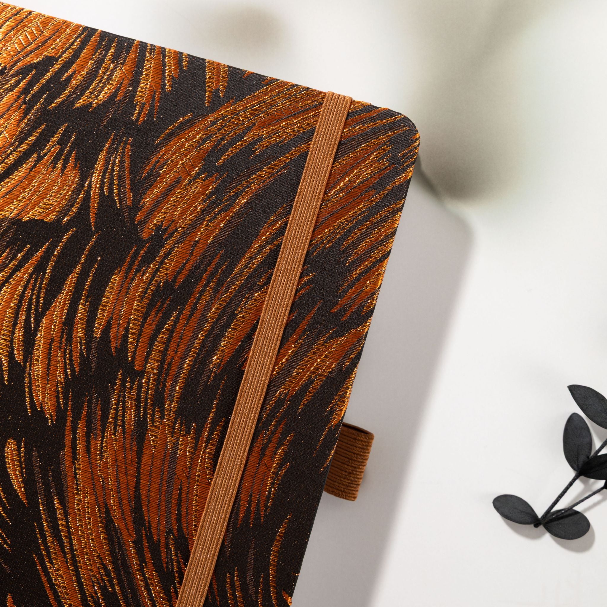 Close-up of the deep brown and copper metallic woven fabric cover with elastic band, placed next to a black leaf stem