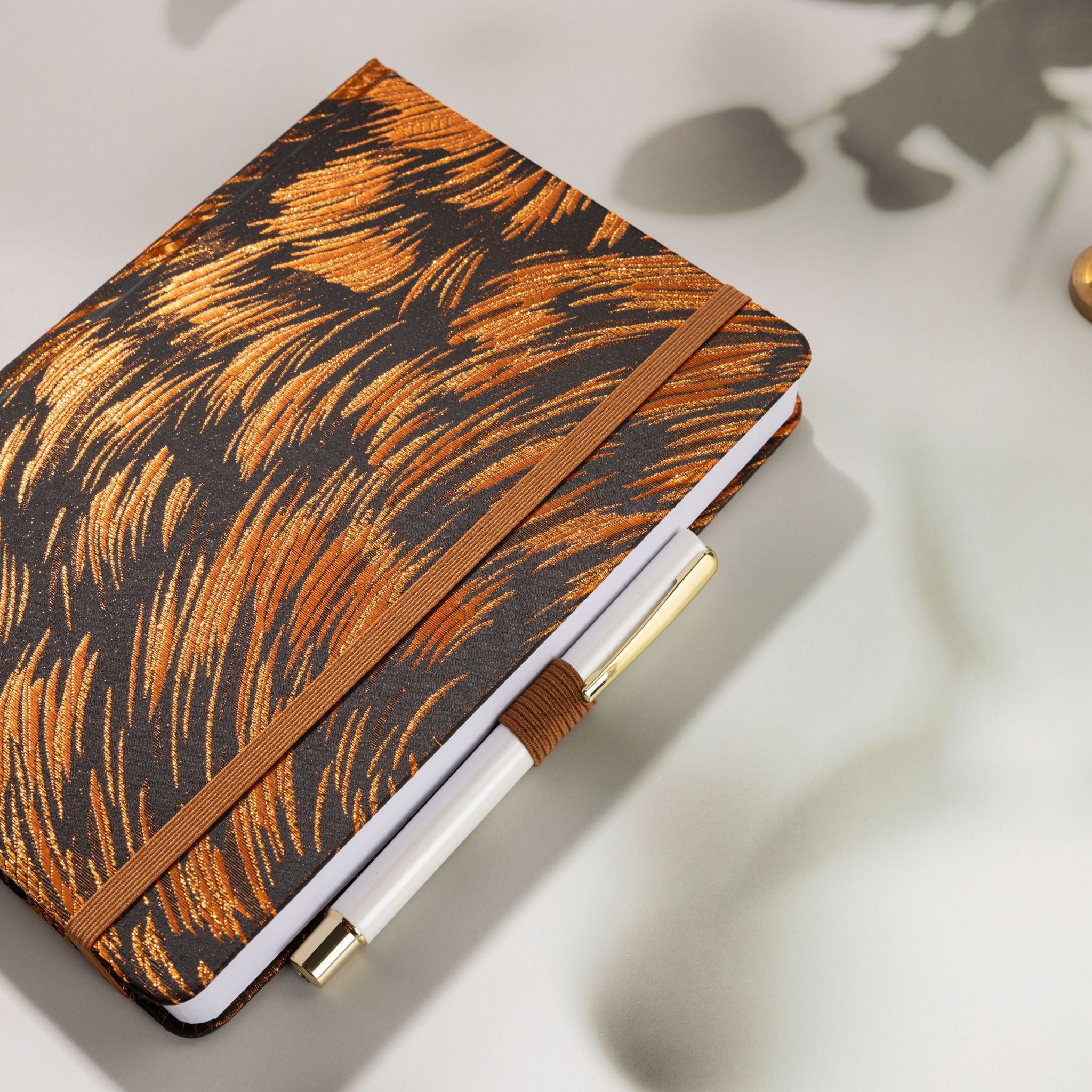 Brown metallic fabric notebook secured with elastic band, holding a white and gold pen in the side loop