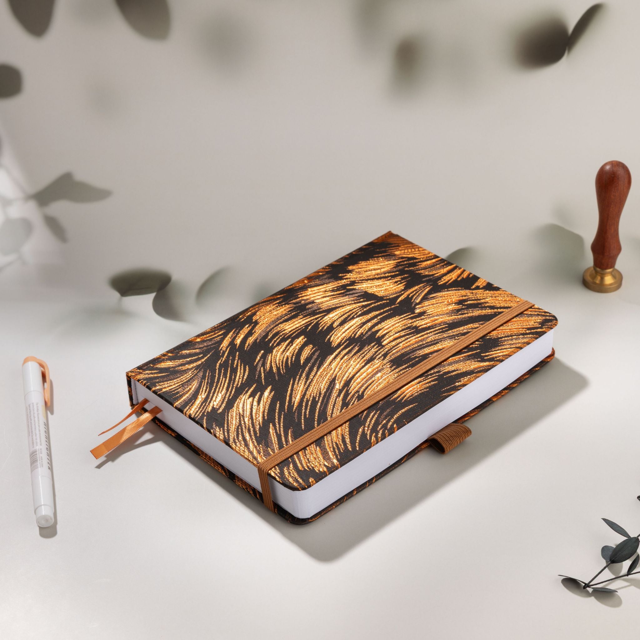 Styled flat lay of the copper brown texture notebook with a white gel pen, wooden wax seal stamp, and botanical shadows on a light surface