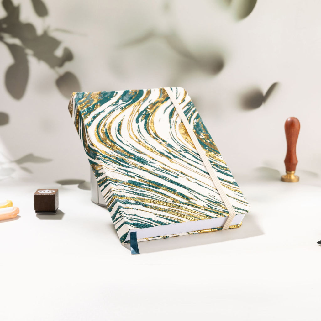 Dark green and gold marble fabric notebook standing upright on a white desk with a wax seal stamp