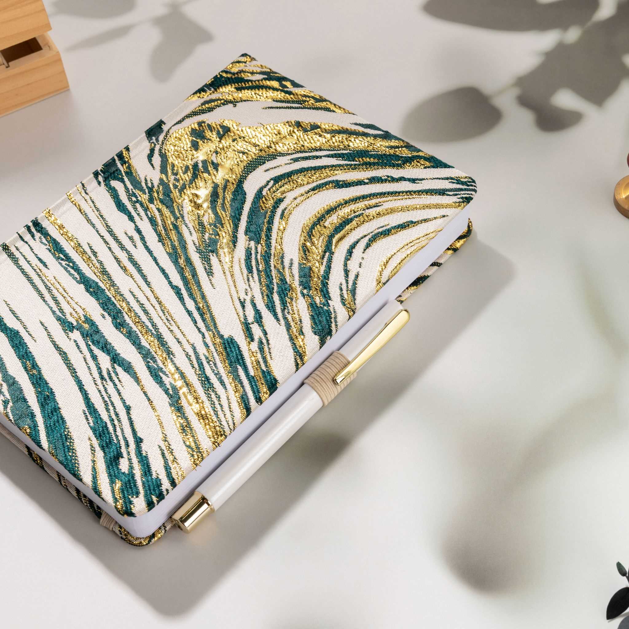 Deep green and gold swirl fabric notebook with elastic band, holding a white and gold pen.