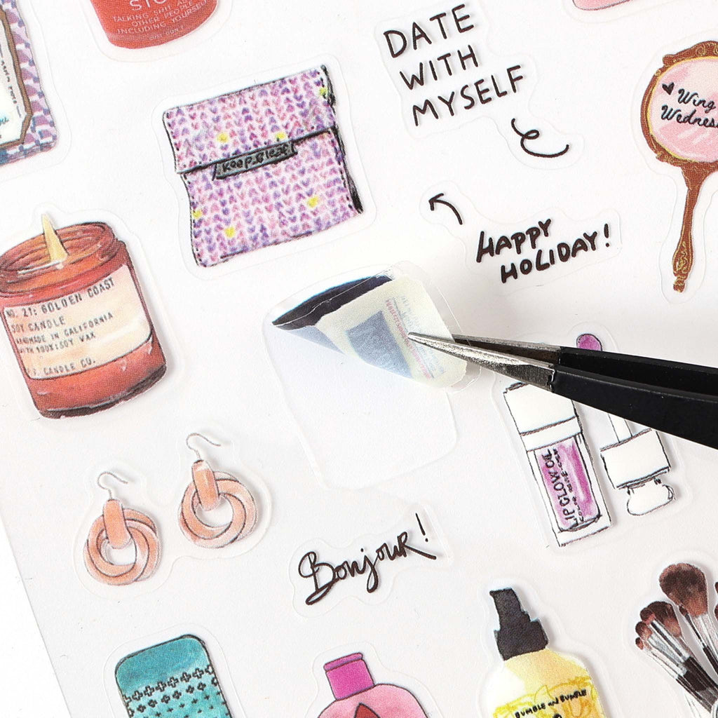 A close-up of a watercolor PET sticker being peeled off its backing using black tweezers, surrounded by illustrated items including a scented candle, a pouch, earrings, lip gloss, and a hand mirror, with handwritten phrases like "DATE WITH MYSELF" and "HAPPY HOLIDAY!".