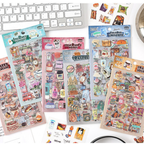 Six Everyday Life watercolor PET sticker themes displayed on a white desk with a keyboard, including Sweet Dreamland, Journey Wonders, Fragrance Notes, Travel List, Colored Universe, and Snack Journal, with a few loose stickers scattered in the foreground.