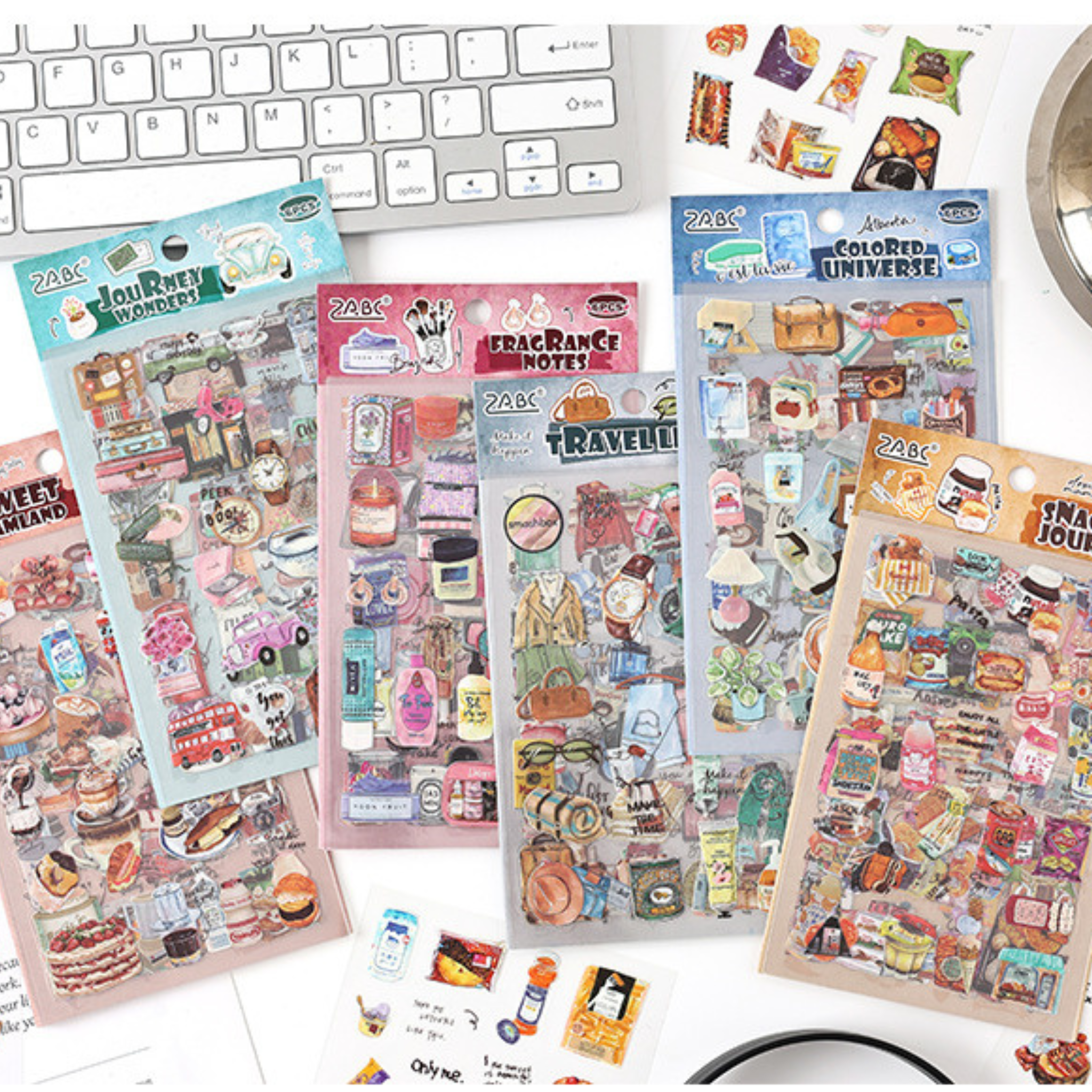 Six Everyday Life watercolor PET sticker themes displayed on a white desk with a keyboard, including Sweet Dreamland, Journey Wonders, Fragrance Notes, Travel List, Colored Universe, and Snack Journal, with a few loose stickers scattered in the foreground.