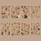 Eight sheets of Autumn Song gold foil PET stickers displayed on a warm beige background, featuring fall-themed botanical elements such as acorns, pinecones, dried leaves, mushrooms, berries, sunflowers, butterflies, and a small hedgehog.
