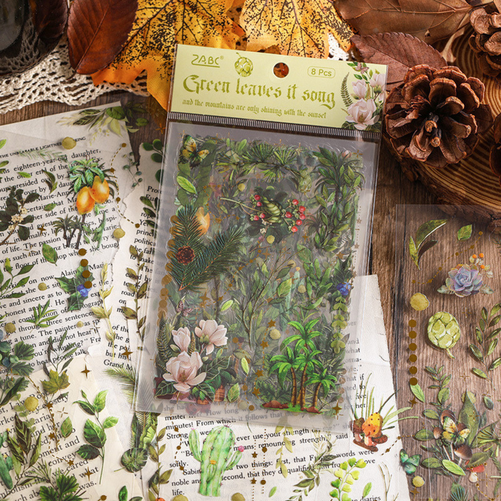 The Green Leaves Song theme from the Forest Song gold foil PET sticker collection, displayed in its hang-hole OPP packaging on a rustic wooden surface with old book pages, pinecones, and dried autumn leaves.
