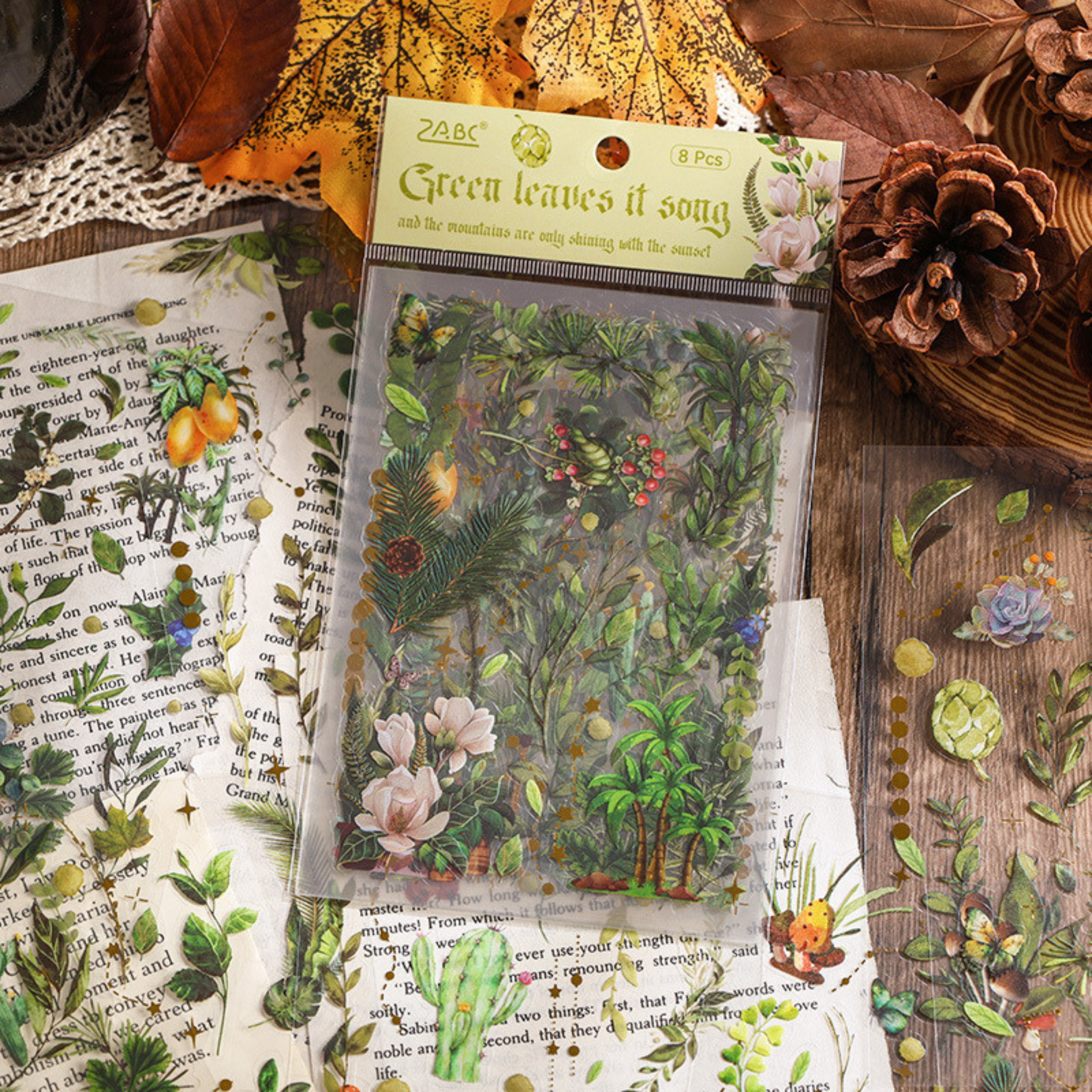 The Green Leaves Song theme from the Forest Song gold foil PET sticker collection, displayed in its hang-hole OPP packaging on a rustic wooden surface with old book pages, pinecones, and dried autumn leaves.