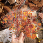 A hand holding several spread-out sheets of the Autumn Song gold foil PET stickers, featuring autumn-themed illustrations including red and orange maple leaves, pumpkins, acorns, pears, a butterfly, and a small squirrel against a rustic wooden background.