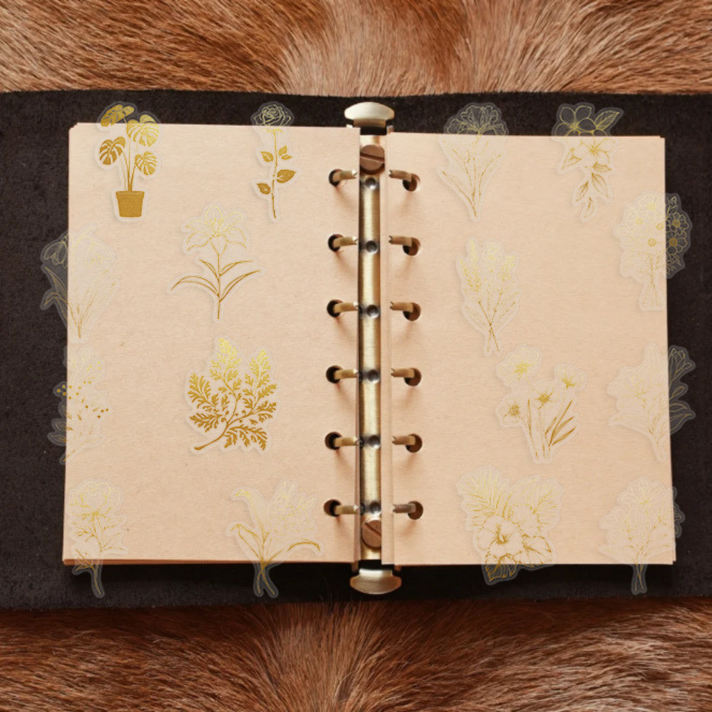 A ring-bound planner opened on a brown surface, decorated with various gold foil floral PET stickers including potted plants, lilies, and roses on cream-colored pages.