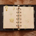 A ring-bound planner opened on a brown surface, decorated with various gold foil floral PET stickers including potted plants, lilies, and roses on cream-colored pages.