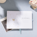 Open notebook featuring gold foil 'M' logo and 'This Journal Belongs To' page on a green scalloped journal