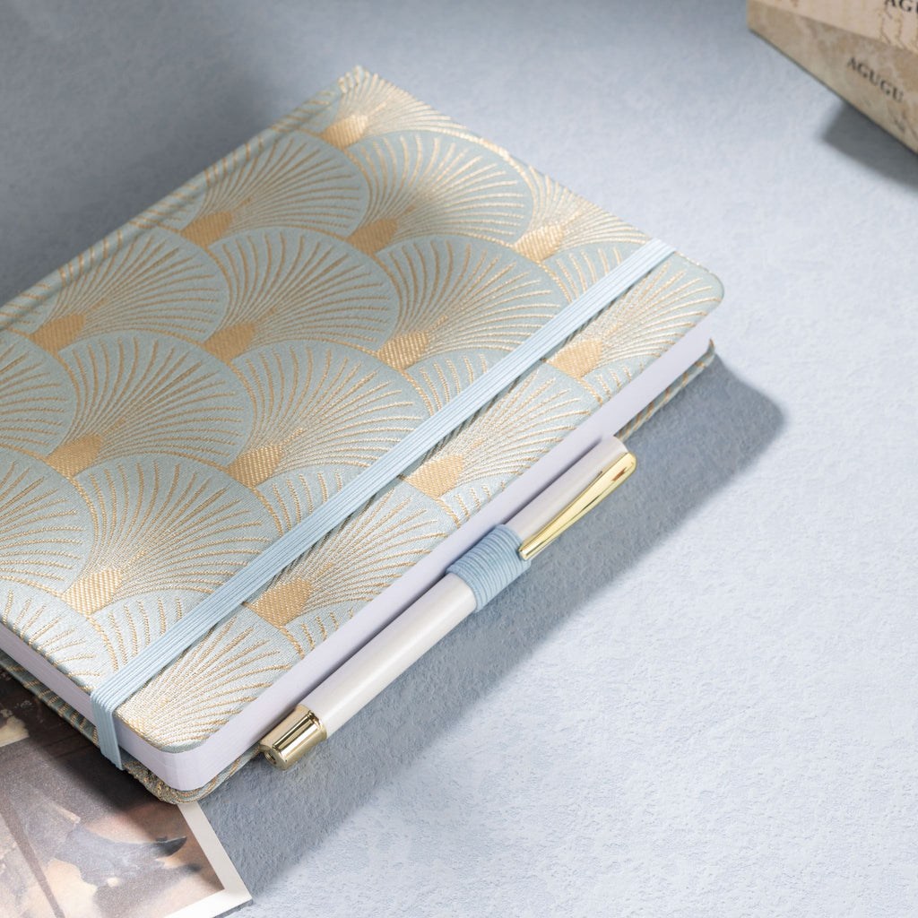 Green and gold woven fabric notebook with matching elastic band, holding a white and gold pen
