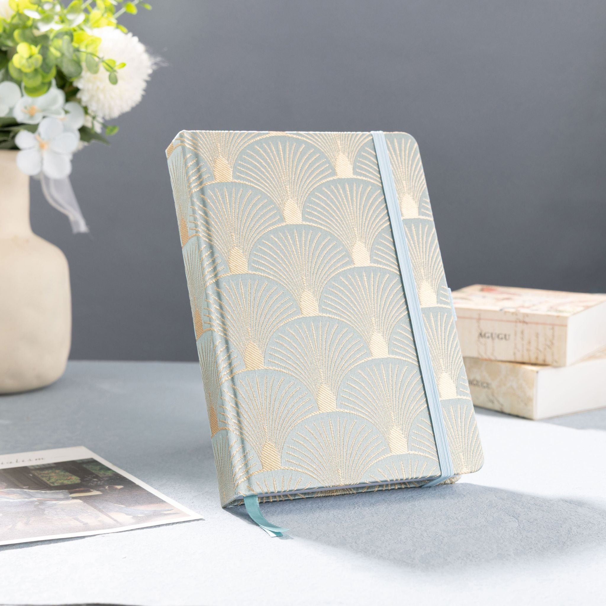 A5 green and gold scalloped fabric notebook standing upright on a gray table with a vase of white flowers