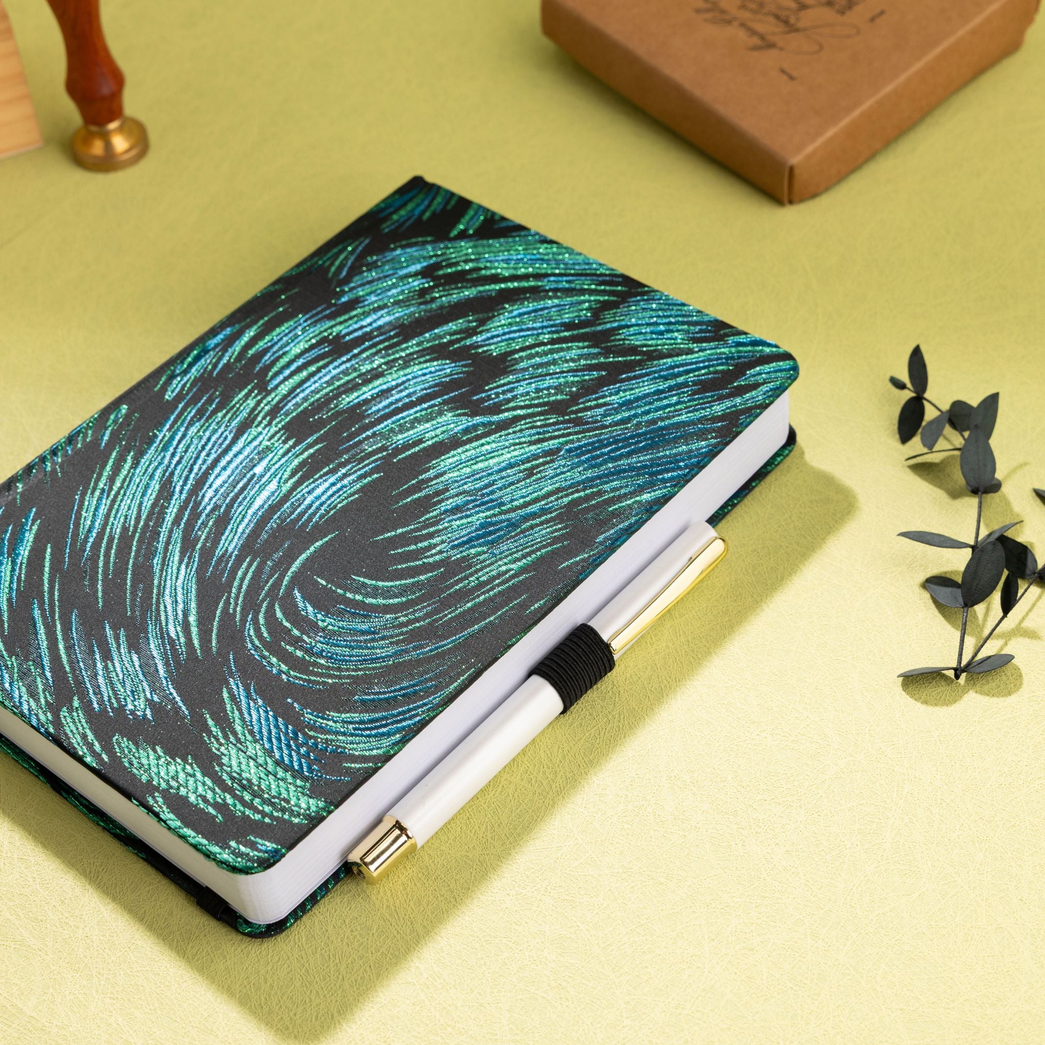 Black and green embroidered fabric notebook with elastic band, holding a white and gold pen