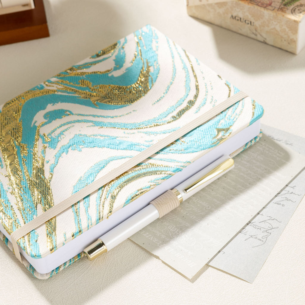 Light blue gold swirl cloth notebook secured with elastic band, holding a gold pen in the loop
