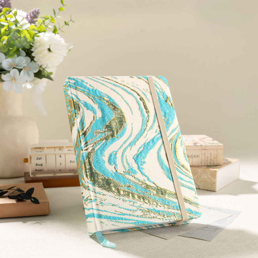 Styled desktop shot of the light blue and gold marble cloth notebook standing upright, accompanied by white flowers and a wooden calendar block in warm sunlight.