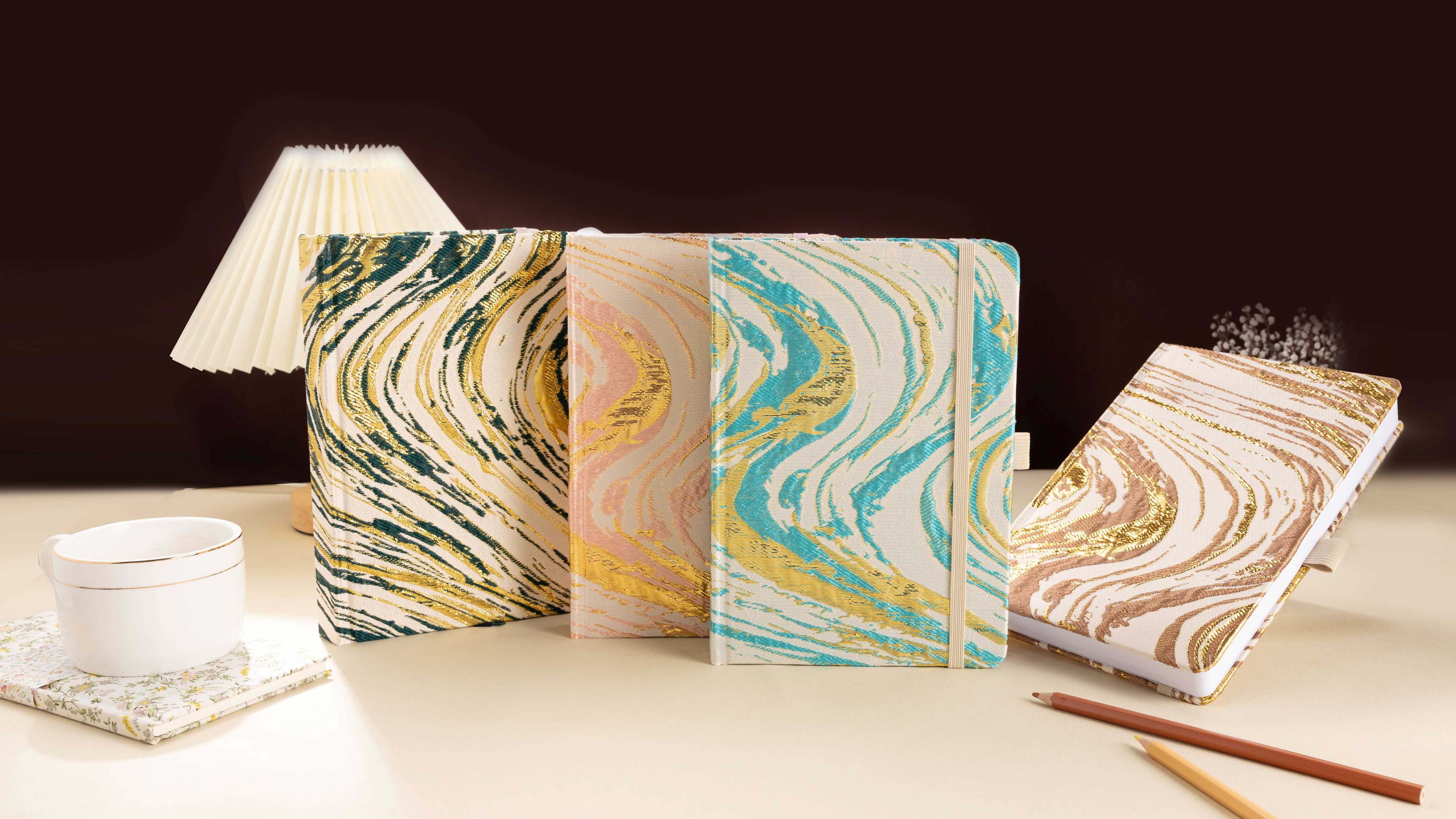 Four A5 woven fabric notebooks with metallic marble swirls in emerald gold, blush pink, teal gold, and bronze brown, arranged on a cream desk with a pleated lamp, coffee cup, and pencils.