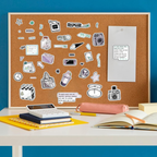 A cork bulletin board covered with assorted die-cut stickers, displayed on a white desk against a bright blue wall. The desk also holds notebooks, a calculator, a pink pencil case, and an open book.
