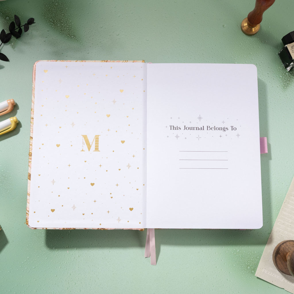 Inside title page featuring gold foil 'M' logo and 'This Journal Belongs To' section on a pink journal