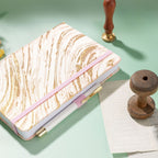 Blush pink gold swirl fabric notebook with elastic band, holding a white and gold pen