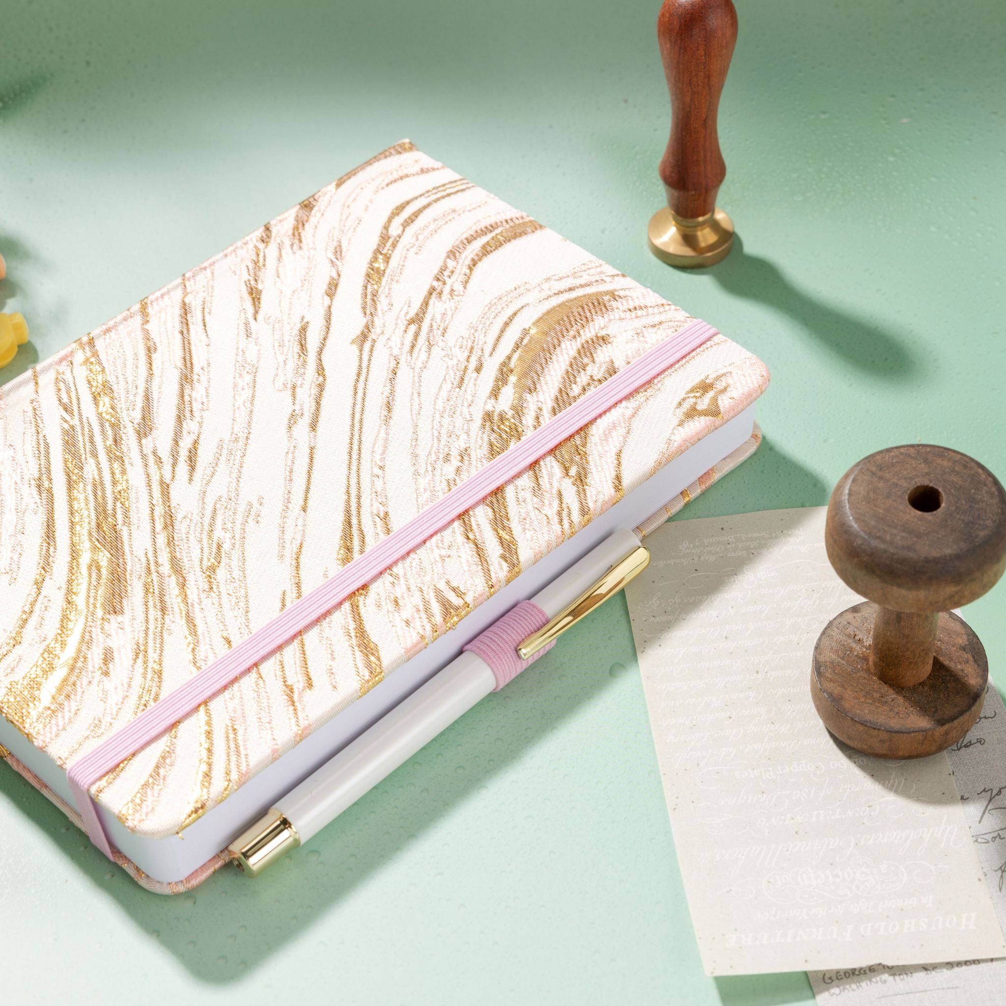 Blush pink gold swirl fabric notebook with elastic band, holding a white and gold pen