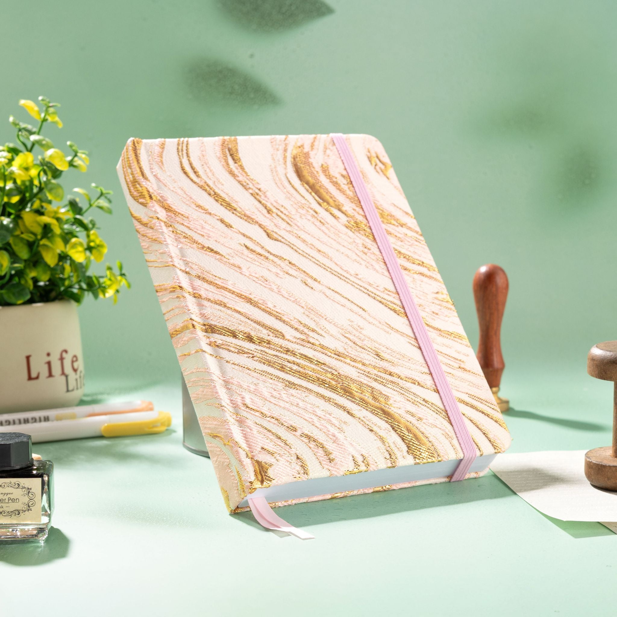 A5 pink and gold marble fabric notebook standing upright on a mint green desk with flowers and a wax seal